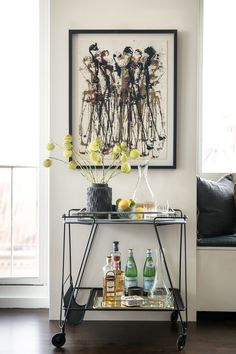 a bar cart with bottles and glasses on it in front of a framed art piece