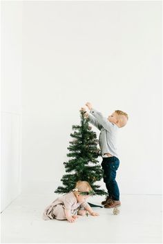 two small children playing with a christmas tree in front of a white wall and floor