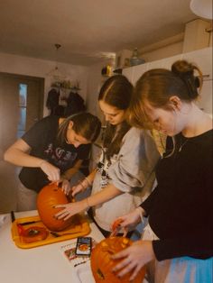 three young women carving pumpkins on top of a kitchen counter with other people around