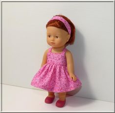 a doll with red hair wearing a pink dress and headband standing against a white wall