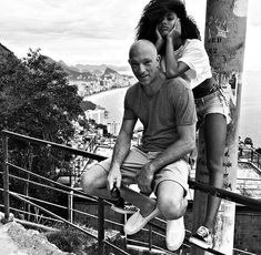 a man sitting on top of a metal rail next to a woman near the ocean