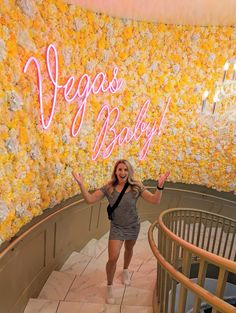 a woman standing in front of a flower wall holding her arms up with both hands