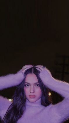 a woman with long dark hair wearing a purple sweater and holding her hands behind her head