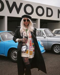 a woman standing in front of a blue car wearing sunglasses and a black coat with patches on it