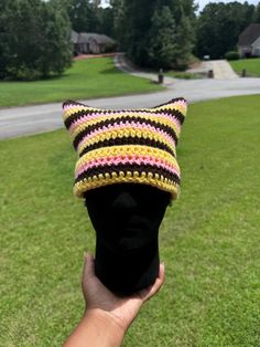 a hand holding up a knitted hat on top of a grass covered field with trees in the background