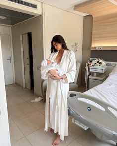 a woman in a white robe holding a baby while standing next to a hospital bed