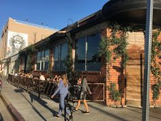 people are walking down the sidewalk in front of a building with vines growing on it