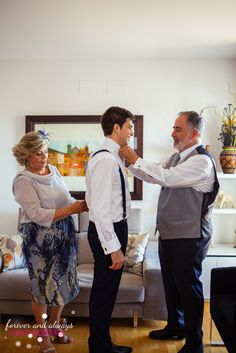 two men and a woman standing in a living room with one adjusting the other's tie