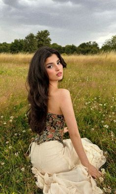 a woman sitting on the ground in a field wearing a dress and looking off into the distance