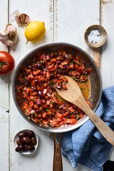 a pan filled with tomatoes and olives next to other ingredients on a white wooden table