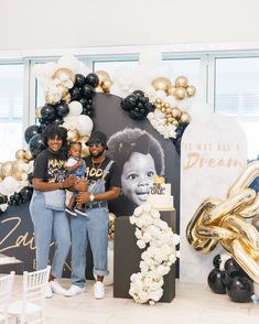 two women standing in front of a backdrop with balloons and flowers, one holding a baby