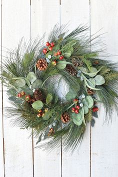 a green wreath with pine cones and berries hanging on a white wooden wall, decorated with greenery