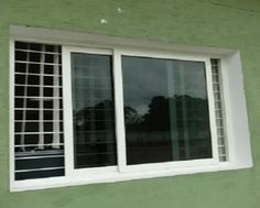 a green building with two windows and white bars on the window sill, reflecting trees in the distance