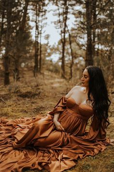 a woman in a brown dress sitting on the ground next to some trees and grass