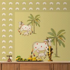 two white cows standing on top of a wooden table next to a wall with palm trees