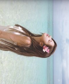 a woman with long brown hair standing next to a pool and holding a flower in her hair