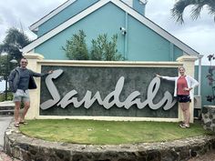 two people standing in front of a sign that says sandels with palm trees behind them