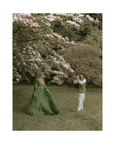 two people standing in the grass with trees behind them and one person taking a photo