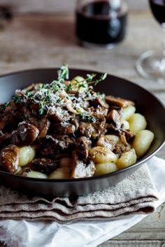 a bowl filled with meat and potatoes on top of a table next to wine glasses