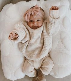 a baby in a white sweater and pink bow laying on top of a pillow with her hands up