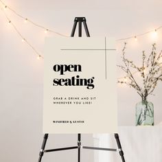 an open seating sign sitting on top of a easel next to a vase with flowers