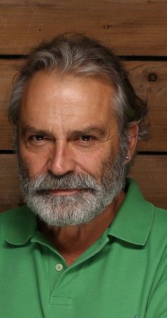 an older man with grey hair and beard wearing a green shirt in front of a wooden wall