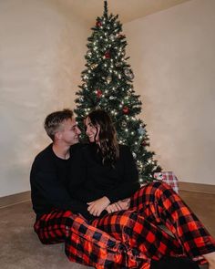 a man and woman are sitting in front of a christmas tree with their arms around each other