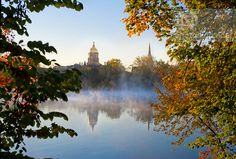 a lake surrounded by trees with fog in the air and a clock tower in the distance