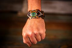 Embrace the fusion of craftsmanship and mystical allure with our Handmade Steampunk Bohemian Leather Cuff Bracelet. This unisex piece is a symphony of details, featuring an intricately carved and painted eyeball and mushrooms, surrounded by geometric patterns inspired by sacred geometry. The design is a bold testament to both artistic vision and spiritual symbolism. Rendered in rich, earthy tones of brown, green, and black, this bracelet interweaves multiple leather strips, creating a complex and textured aesthetic. The antique brass buckle closure adds a vintage touch while ensuring a comfortable fit for wrist sizes ranging from 7 to 9.25 inches. Perfect for festivals like Burning Man, spiritual gatherings, or everyday wear, this leather cuff serves as more than just an accessory—it's a p Adjustable Unique Leather Bracelet For Festivals, Festival Leather Bracelet, Unique Hand Wrapped Leather Bracelet For Festivals, Steampunk Adjustable Bracelets For Festivals, Adjustable Steampunk Festival Bracelets, Adjustable Steampunk Bracelets For Festivals, Hand Wrapped Spiritual Leather Bracelet For Festival, Spiritual Hand Wrapped Leather Bracelet For Festivals, Handmade Adjustable Steampunk Bracelets