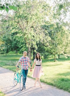 an engaged couple holding hands while walking their bike down a path in the park together