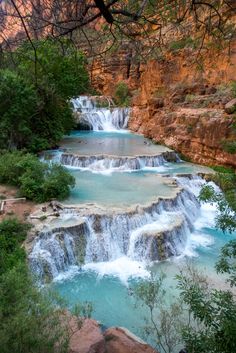 two people are floating in the water near a waterfall and some red rocks with trees