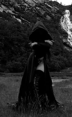 a woman in black clothing and boots is standing near a waterfall with her arms wrapped around her body