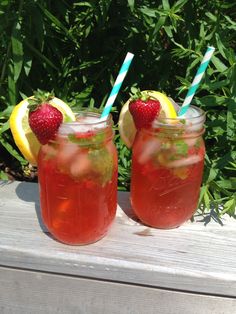 two mason jars with strawberries and lemons sit on a wooden table in front of bushes