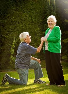 an older man and woman holding hands while sitting on the grass in front of trees