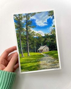 a person holding up a card with a painting of a house in the woods