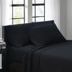 a bed with black sheets and pillows in front of a large window, next to a night stand