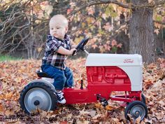 a young boy riding on top of a red toy tractor in the leaves with trees behind him