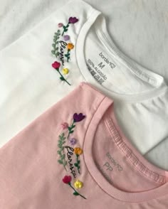 three t - shirts with embroidered flowers on them sitting on a white bedding sheet