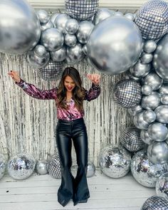 a woman standing in front of silver balloons and disco ball backdrop with her hands up