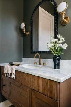 a bathroom vanity with marble counter top and wooden cabinet, gold faucet, white flowers in black vase