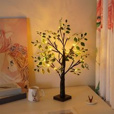 a lighted tree is sitting on a desk next to a book and coffee mugs