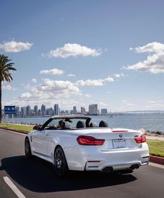 a white convertible car driving down the road next to water and cityscape in the background