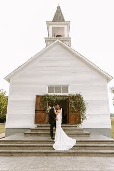 a bride and groom standing in front of a white church with steps leading up to it