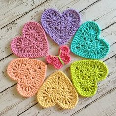 six crocheted hearts are arranged on a white wooden surface, each with different colors