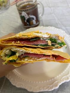 a hand holding a sandwich on top of a white plate next to a cup and saucer