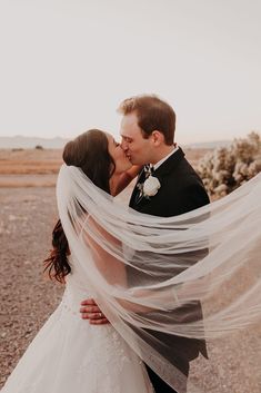 a bride and groom kissing in the desert with veil blowing over their heads as they stand close to each other