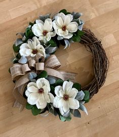 a wreath with white flowers and green leaves on a wooden floor next to a brown ribbon