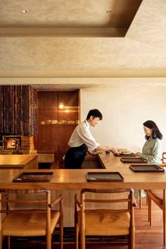 a man and woman standing at a table in a room with wooden chairs around it