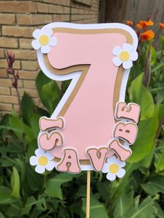 a pink number one cake topper with daisies in front of some plants and flowers