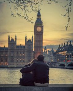 two people sitting on a bench in front of the water with a clock tower in the background